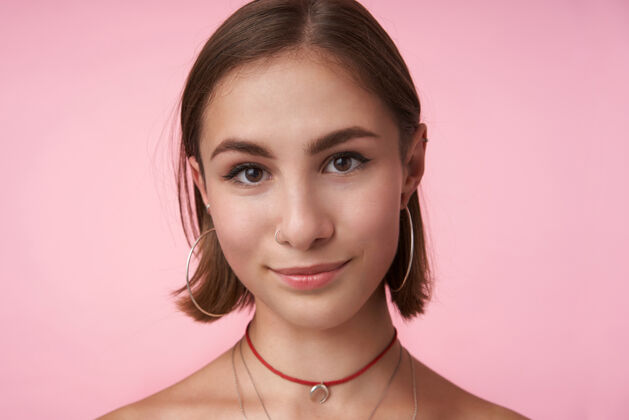 自然年轻漂亮的棕色头发的女人的肖像 自然的妆容 正面带着愉快的微笑 穿着时髦的配饰 站在粉红色的墙上休闲发型耳环