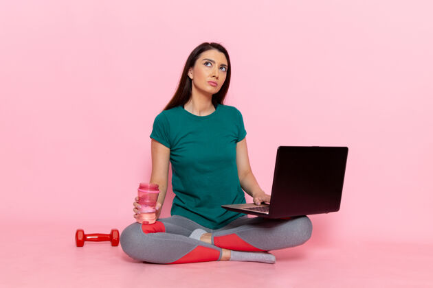 坐着正面图身着绿色t恤的年轻女性在粉色墙上的笔记本电脑上工作腰部运动锻炼美丽苗条女性运动年轻人电脑女性