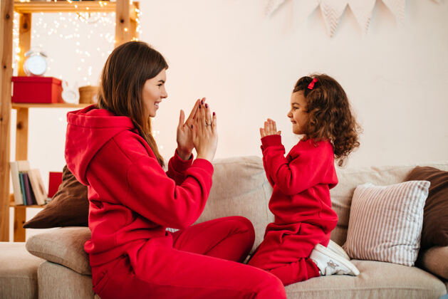 享受快乐的年轻母亲和女儿在沙发上玩耍室内拍摄迷人的妈妈和穿着红色服装的未成年儿童公寓房子妈妈