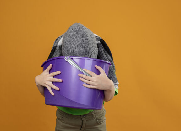 站着生病的小男孩穿着绿色t恤 戴着暖和的围巾 戴着帽子 手里拿着垃圾 站在橙色的墙上感到恶心温暖垃圾抱着