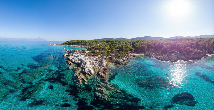 观点广角拍摄的爱琴海海岸 蓝色透明的海水 周围的绿色植物 岩石 灌木丛和树木 小山 帕莫拉马从无人机 希腊的看法海岸海湾度假村