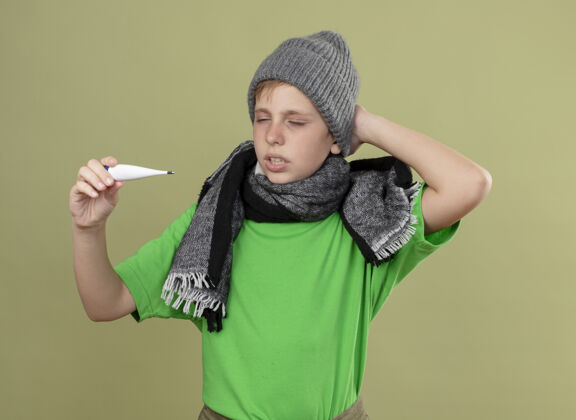 看生病的小男孩穿着绿色t恤 戴着暖和的围巾 戴着帽子 感觉不舒服 拿着体温计 困惑地看着它 站在明亮的墙上感觉抱着男孩