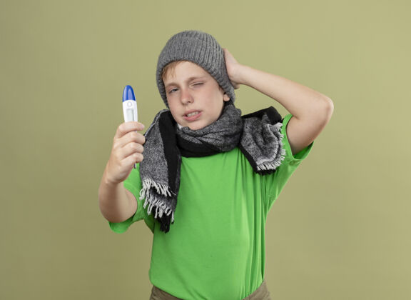 看生病的小男孩穿着绿色t恤 戴着暖和的围巾 戴着帽子 站在明亮的墙上感到不舒服显示温度计光