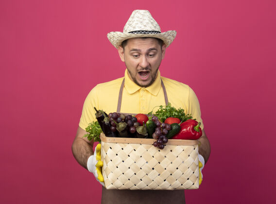 男年轻的园丁穿着连体衣 戴着帽子 戴着工作手套 手里拿着装满蔬菜的箱子 站在粉色的墙上 看上去既惊讶又惊讶花园工人男人
