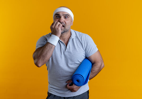 运动成熟的运动型男人戴着头巾 拿着瑜伽垫 站在橙色的墙上 紧张而紧张紧张抱着人