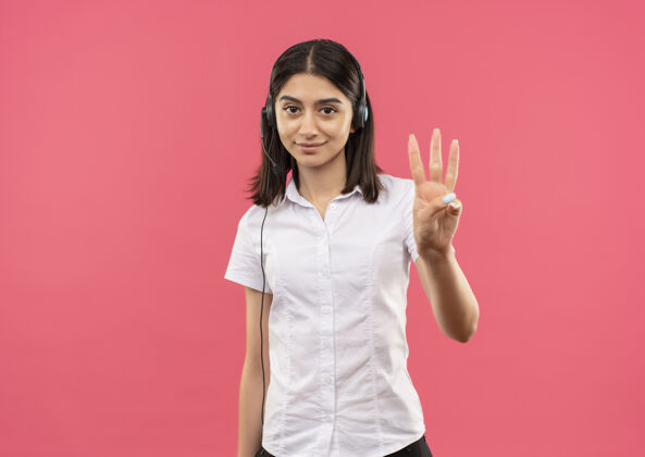年轻的穿着白衬衫戴着耳机的年轻女孩 手指着站在粉色墙上微笑的三号女孩号码微笑指着