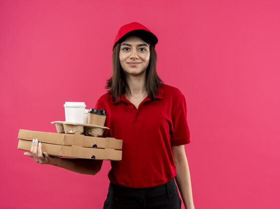 食物身穿红色马球衫 头戴帽子 拿着披萨盒和食品包的年轻送货女孩站在粉红色的墙上 脸上洋溢着幸福的笑容盒子送货穿