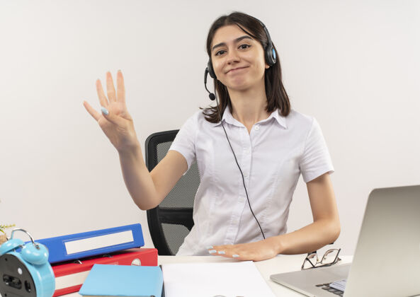 人穿着白衬衫 戴着耳机的年轻女孩 三号手指微笑着坐在桌旁 文件夹和笔记本电脑盖在白墙上桌子女性手势