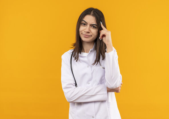 办公室身穿白大褂 脖子上戴着听诊器的年轻女医生站在橙色的墙上 带着怀疑的表情望着前方女人年轻人外套