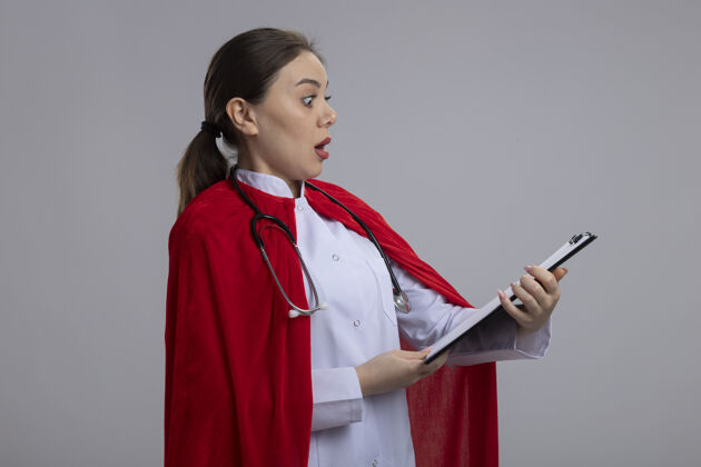 看穿着白色医疗制服 穿着红色超级英雄斗篷 手持听诊器的女医生站在白色的墙上 展示着空白页的剪贴板 看上去既惊讶又惊讶剪贴板人医疗