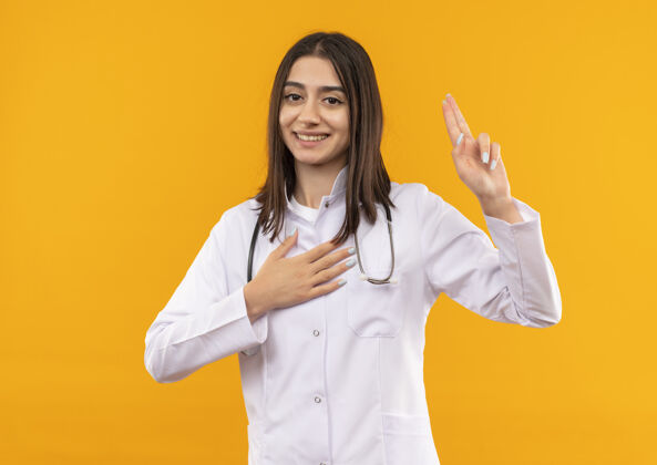 姿势身穿白大褂 脖子上戴着听诊器的年轻女医生站在橘色的墙上向前方宣誓姿势手势女人