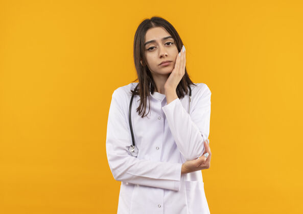 女人身穿白大褂 脖子上戴着听诊器的年轻女医生站在橘色的墙上 带着沉思的表情望着前方年轻人女人脖子