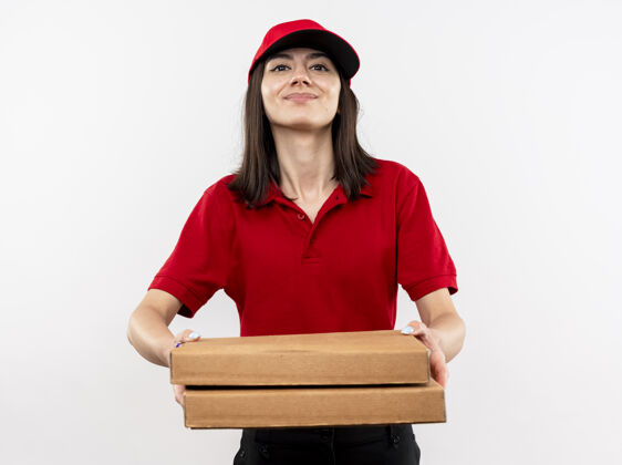 制服年轻的送货员身穿红色制服 戴着帽子 手里拿着披萨盒 站在白色的墙上 脸上洋溢着幸福的笑容女孩站着抱着