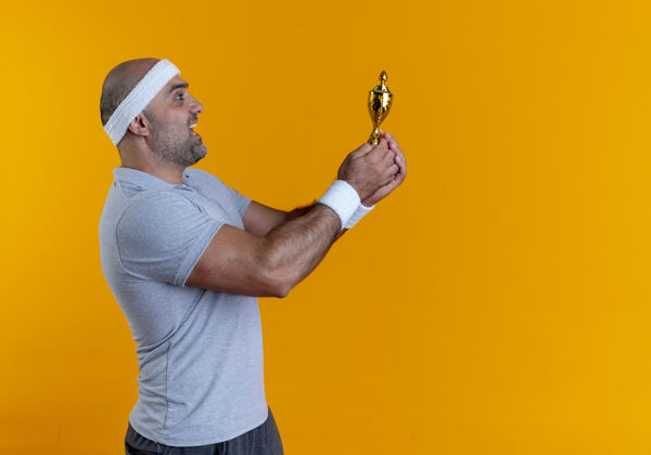 抱戴着头巾的成熟的运动型男人手持奖杯快乐而兴奋地站在橙色的墙上市民成熟兴奋