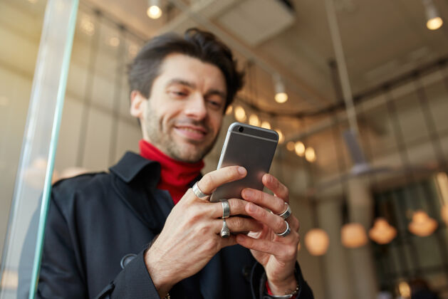 男性举着戒指的男人的手的特写保持手机 同时在城市咖啡馆背景上摆姿势 给朋友发短信 微笑着 同时积极地看着屏幕胡须咖啡馆黑发