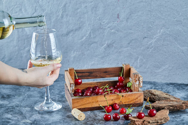 木头一个女人一边倒白葡萄酒 一边拿着一个玻璃杯和一盒樱桃放在蓝色的表面上盒子浆果饮料