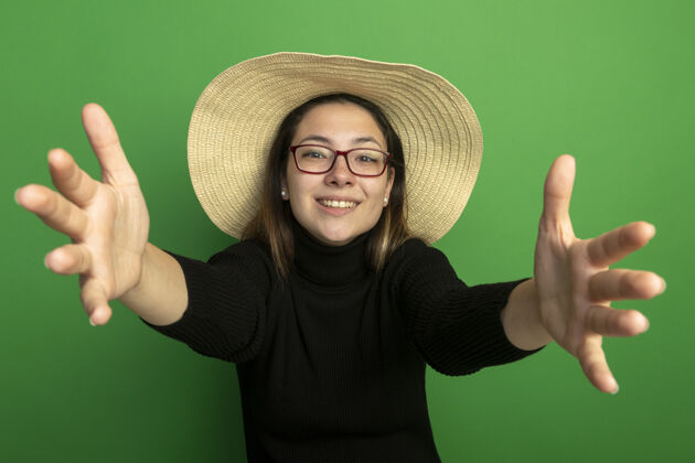 制作年轻漂亮的女人戴着夏天的帽子 穿着黑色高领毛衣 戴着眼镜 做着欢迎的手势 开心而积极的微笑友好地站在绿色的墙上美丽年轻帽子