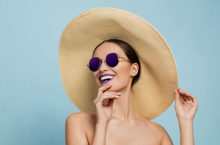护理美丽女人的肖像 明亮的妆容 蓝色工作室背景上的帽子和太阳镜时髦和时尚的制作和发型夏天的颜色美丽 时尚和广告概念笑保持漂亮新鲜