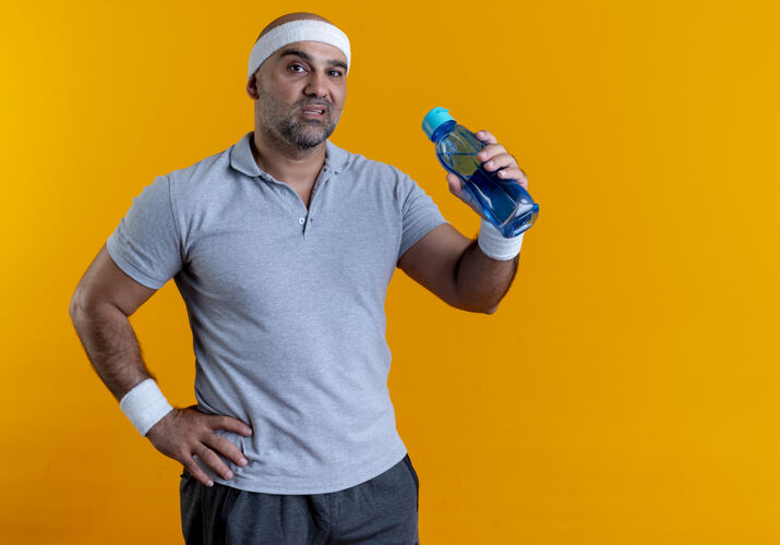 脸戴着头巾的成熟的运动型男人拿着一瓶水望着前面 严肃的脸站在橙色的墙上男水运动
