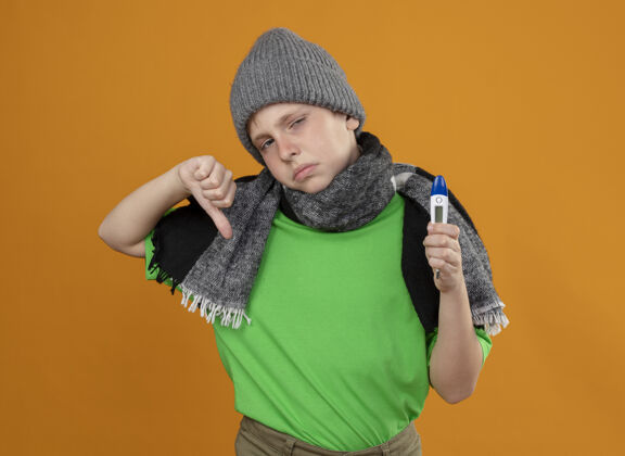 按住生病的小男孩穿着绿色t恤 戴着暖和的围巾 戴着帽子 拿着温度计 感觉不舒服 生病 不开心 站在橙色的墙上竖起大拇指男孩帽子不开心