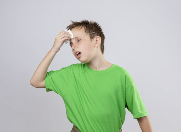 感情生病的小男孩穿着绿色t恤 站在白色的墙上用纸巾擦着脑袋 感觉不舒服男孩立场餐巾