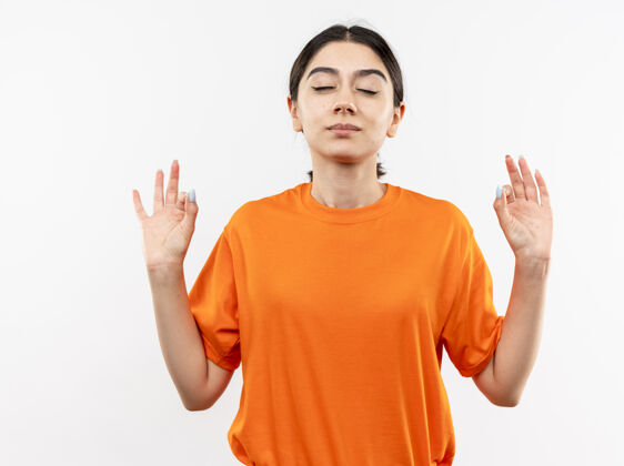 佩戴穿着橙色t恤的年轻女孩闭着眼睛放松 手指站在白墙上做冥想手势制造闭合女孩