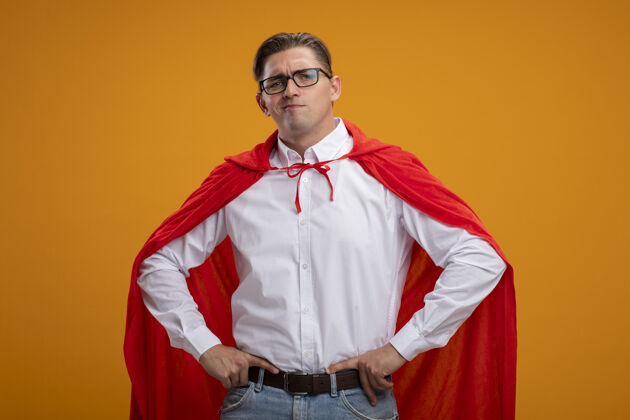 眼镜超级英雄商人穿着红色斗篷 戴着眼镜 严肃的脸 双臂放在臀部 站在橙色的墙上站臀部超级英雄