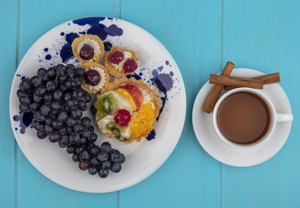 盘子盘子上的黑葡萄顶视图 蓝色木制背景上有一杯茶和肉桂棒杯子茶肉桂
