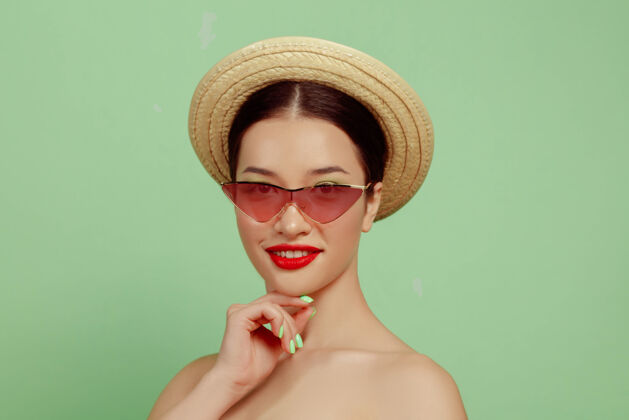 年轻美丽女人的肖像 明亮的妆容 红色的眼镜和帽子 绿色的工作室背景时尚和时尚的制作和发型夏天的颜色美丽 时尚和广告概念摆姿势封闭头新鲜