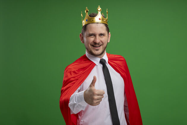 站立身穿红色斗篷的超级英雄商人戴着王冠 微笑着 高举大拇指站在绿色的墙上拇指穿超级英雄
