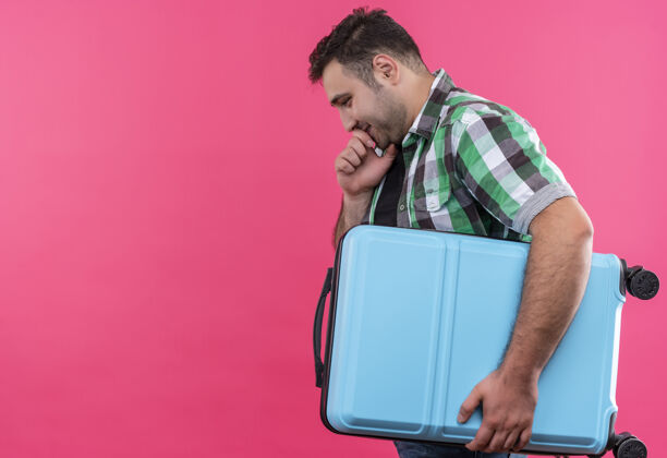 抱着穿着格子衬衫的年轻旅行家手里拿着手提箱 脸上挂着沉思的表情 站在粉红色的墙上手提箱男人检查