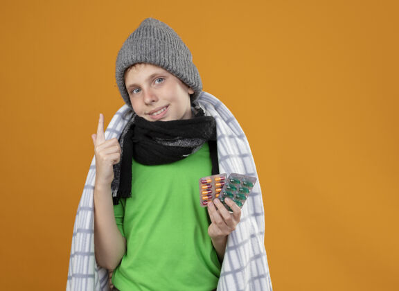 更好生病的小男孩戴着暖和的帽子 围着围巾裹在毯子里 展示着药片 感觉好极了 微笑着 食指站在橙色的墙上温暖目录看