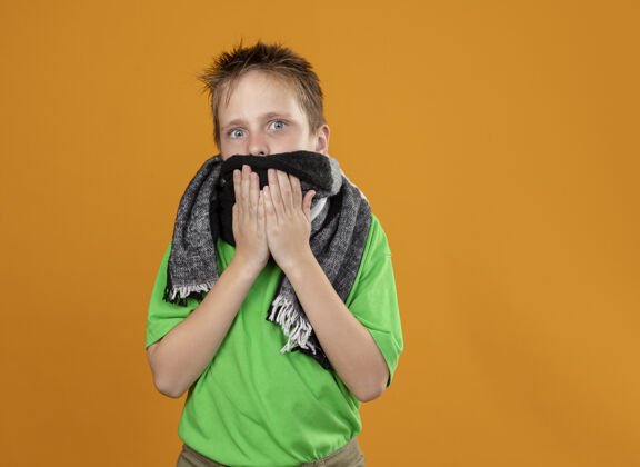 温暖生病的小男孩穿着绿色t恤 围着温暖的围巾站在橙色的墙上 看起来很担心看疾病橙色