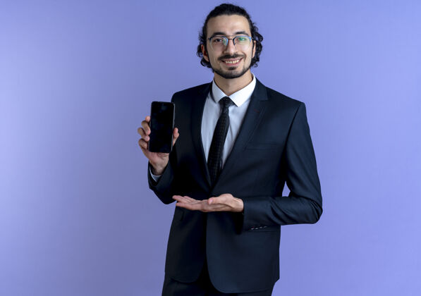 手一位身穿黑色西装 戴着眼镜的商务人士站在蓝色的墙上 展示着智能手机 他的手臂微笑着自信地展示着它商务西装展示