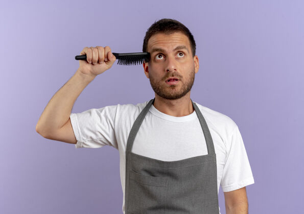 工作围裙上的理发师拿着理发机和发刷站在紫色的墙上人人围裙