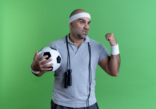 姿势戴着头巾的成熟的运动型男人举着足球举起拳头 严肃的脸站在绿色的墙上向一边看教练人男