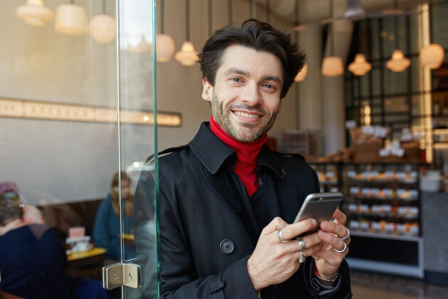 男性室内照片年轻迷人的棕色头发的男性与时尚的发型正面看着相机 面带微笑 而站在咖啡馆内部与手机风衣微笑正面