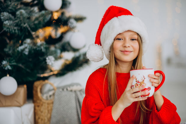 可爱在圣诞树旁喝可可的女孩礼物看时尚