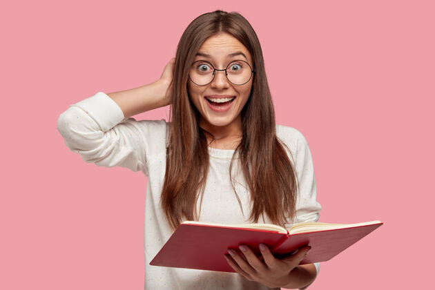 个人漂亮的微笑女孩有着快乐的表情 手放在脑后 拿着红色的课本 微笑着阅读必要的信息 隔着粉红色的墙壁 戴着圆眼镜积极大学教科书