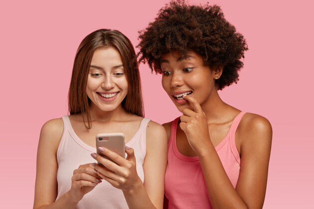 种族室内拍摄好奇开朗的rwo多民族女性通过手机在互联网上阅读新闻 网上购物 站得很近 有跨种族的关系 隔着粉色的墙与世隔绝信息好奇友谊