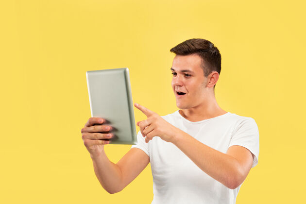 面部黄色工作室背景上的白人年轻人半身肖像穿着衬衫的漂亮男模人类情感的概念 面部表情 销售 广告使用平板电脑 看起来很开心事业商人表情