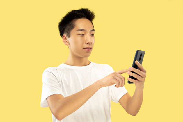 成功黄色工作室背景上的韩国年轻人半身像穿白衬衫的男模使用智能手机人类情感的概念 面部表情正面视图流行色彩衬衫工作员工
