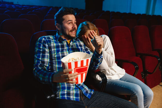 电影院迷人的年轻白种人夫妇在电影院 房子或电影院看电影看起来富于表现力 惊讶和情绪化独自坐着玩关系 爱情 家庭 周末时间娱乐座位舒适