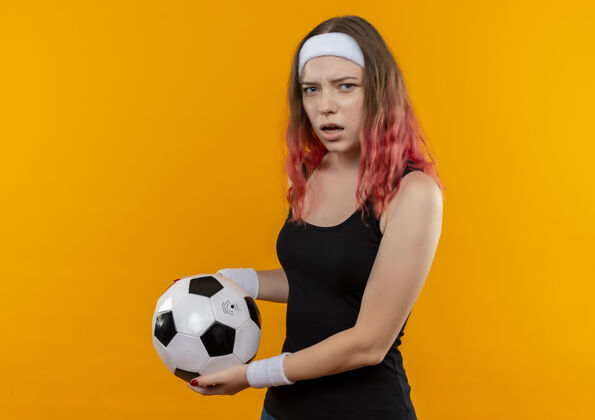 足球身穿运动服的年轻健身女士站在橙色的墙上 手里拿着足球 既不高兴又困惑年轻看健身