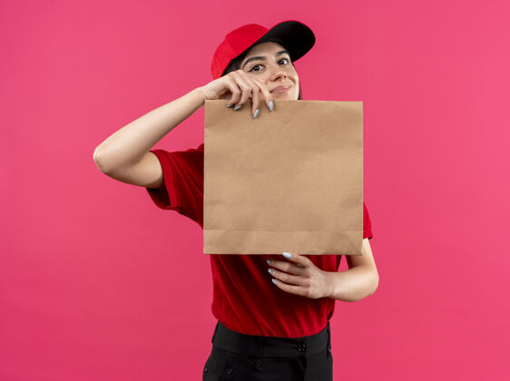 女孩穿着红色马球衫 戴着帽子的年轻送货女孩拿着纸包 面带微笑地看着相机站在粉色背景下包装年轻立场
