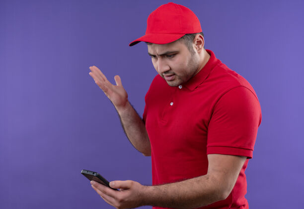 人年轻的送货员穿着红色制服 戴着帽子 站在紫色的路边 看着智能手机的屏幕 既不高兴又失望屏幕站不高兴