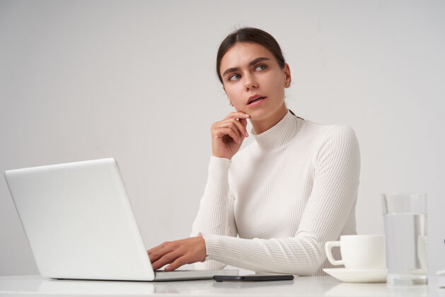 Poloneck迷惑不解的年轻美丽的黑发女士 自然的妆容 用抬起的手抚摸着她的脸 若有所思地看着 在现代化的办公室里工作 笔记本电脑在白色的墙上电话正式思想