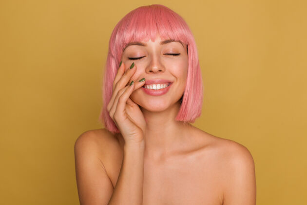 头发可爱漂亮的年轻粉红头发女性 自然妆容 举手轻抚脸庞 在芥末墙上摆姿势时 面带微笑肖像情感美甲