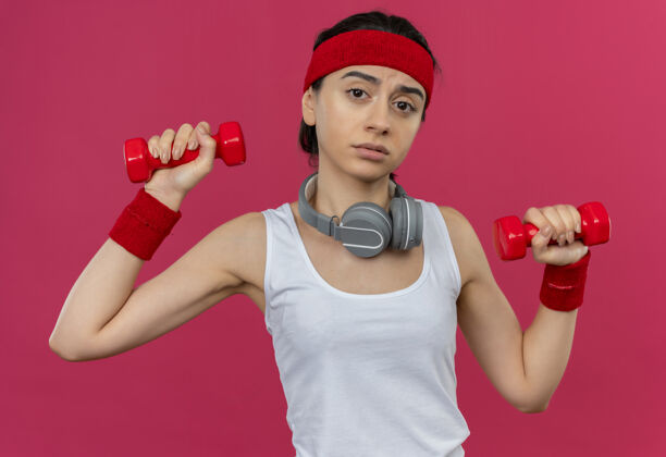 运动身穿运动服的年轻健身女 头上绑着两个哑铃 站在粉红色的墙上 脸上带着悲伤的表情做着运动市民表情健身
