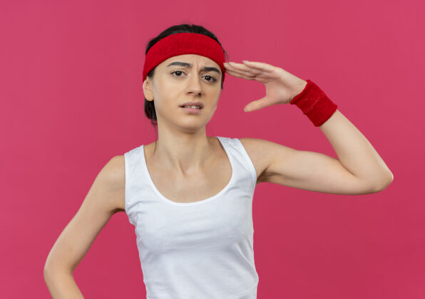 运动员身穿运动服的年轻健身女 头上带着怀疑的表情 站在粉色的墙上敬礼健身表情姿势